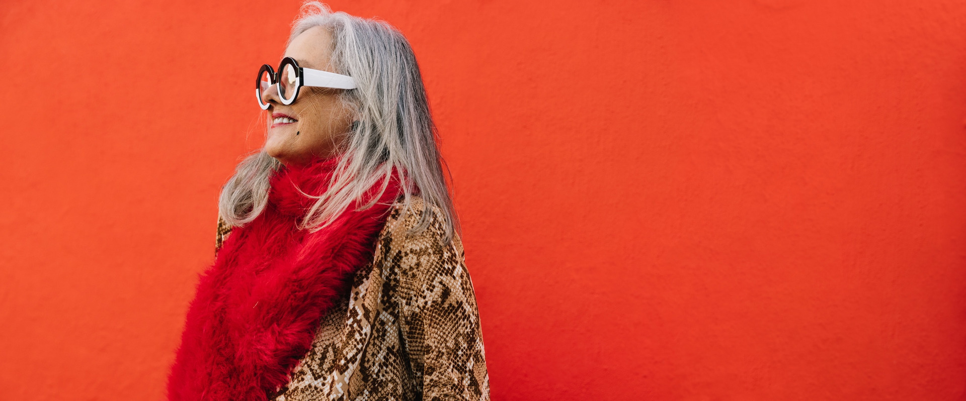 Im Mittelpunkt steht eine ältere Frau mit langen grauen Haaren und runder Brille, die einen roten Schal und eine braun-beige Jacke mit Schlangenmuster trägt. Sie lächelt und schaut zur Seite vor einem leuchtend roten Hintergrund.