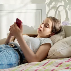 Jugendliche liegt entspannt auf einem Bett und schaut auf ihr rotes Smartphone. 