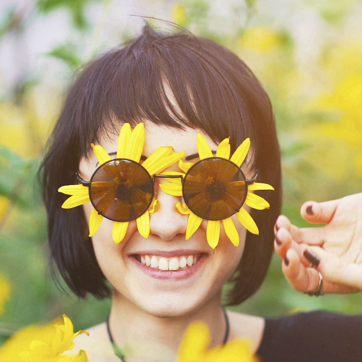 Eine Frau steht in einem Feld gelber Blumen, auch unter ihrer Sonnenbrille befinden sich zwei gelbe Blüten; sie lächelt uns an.