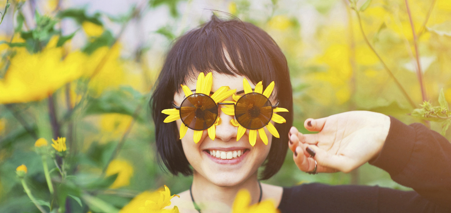 Eine Frau steht in einem Feld gelber Blumen, auch unter ihrer Sonnenbrille befinden sich zwei gelbe Blüten; sie lächelt uns an.