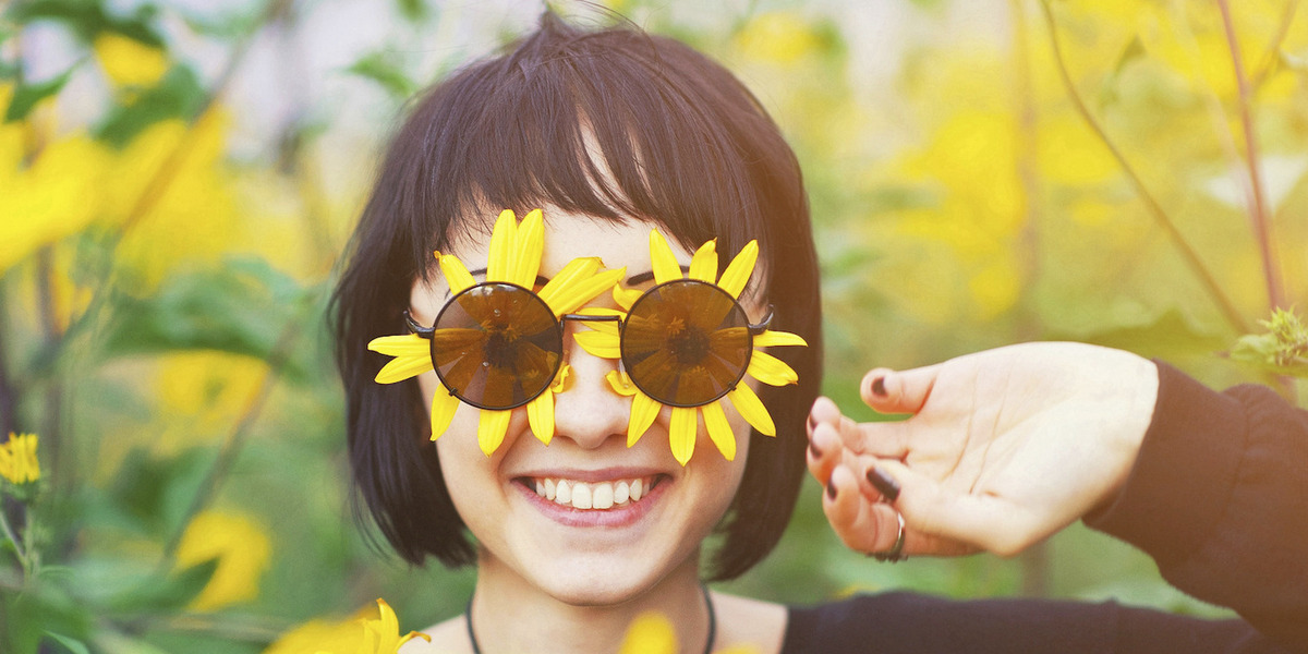Eine Frau steht in einem Feld gelber Blumen, auch unter ihrer Sonnenbrille befinden sich zwei gelbe Blüten; sie lächelt uns an.