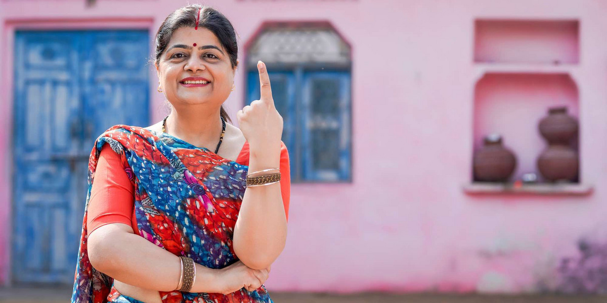 Eine indische Frau mit erhobenem Zeigefinger steht vor einem rosa Gebäude; in Indien wählt man mit dem Fingerabdruck des Zeigefingers