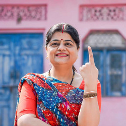 Eine indische Frau mit erhobenem Zeigefinger steht vor einem rosa Gebäude; in Indien wählt man mit dem Fingerabdruck des Zeigefingers