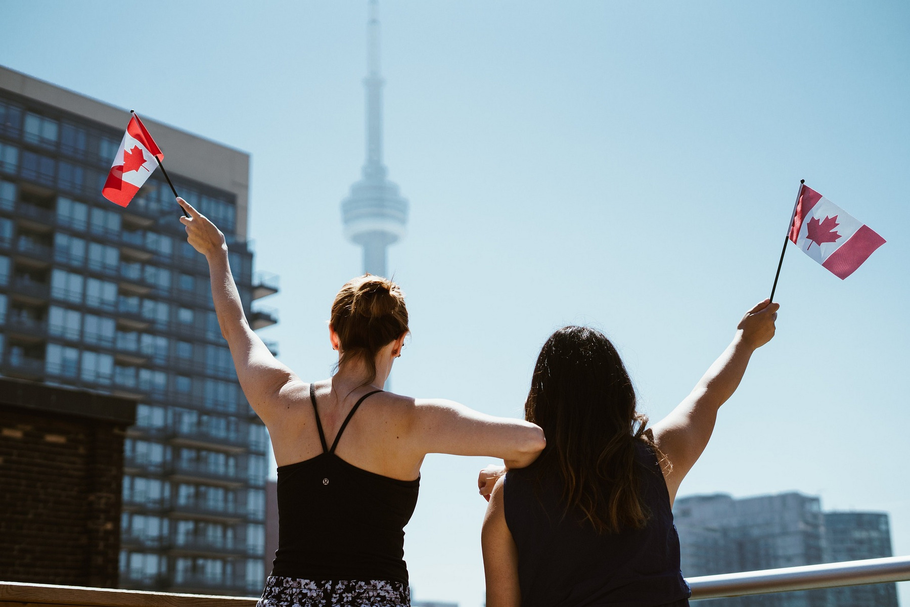 Zwei Frauen stehen mit dem Rücken zur Kamera, vor der Skyline von Toronto, und schwenken kleine kanadische Flaggen.