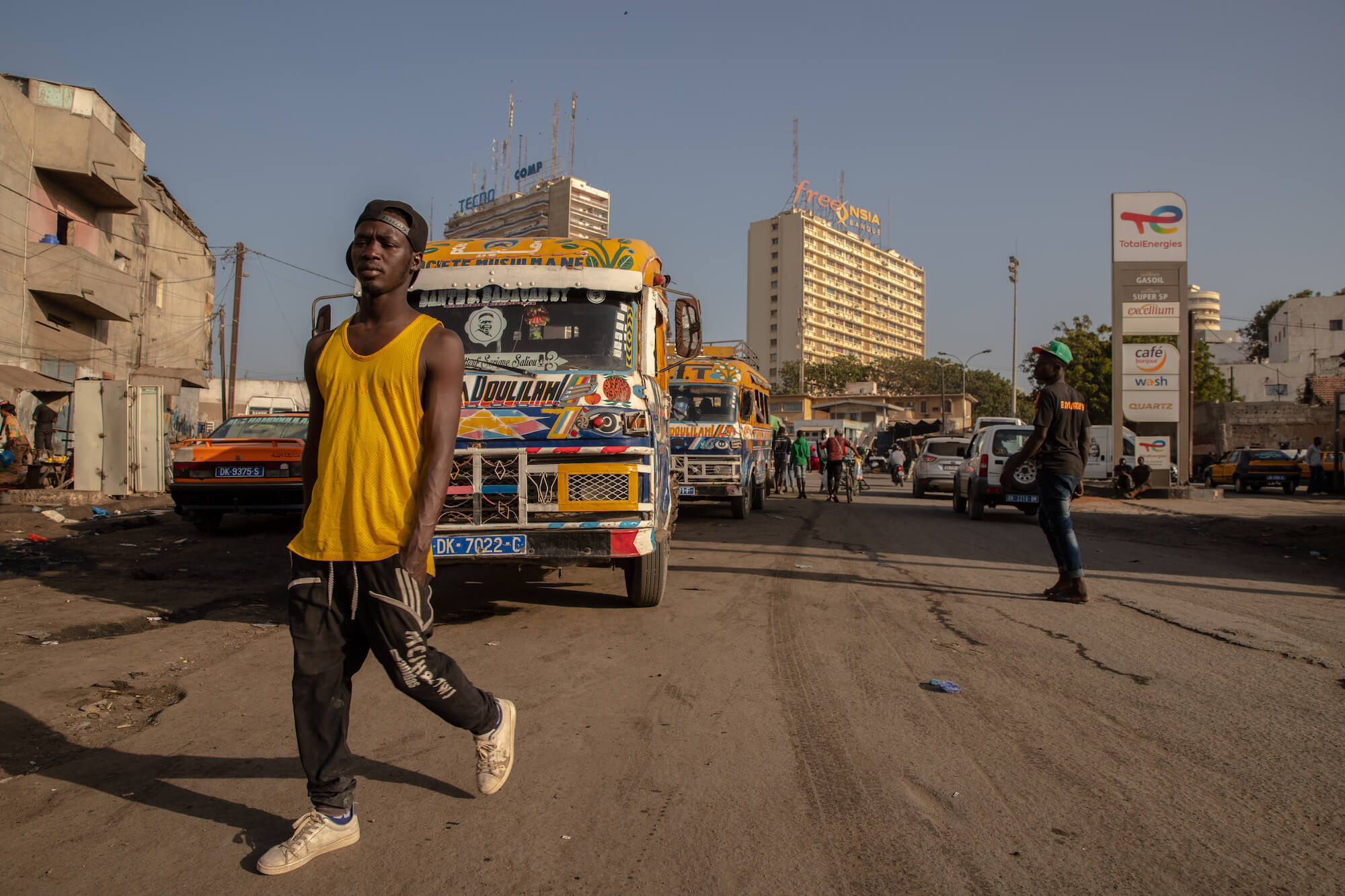 Eine lebendige Straße im Senegal: Im Vordergrund überquert ein Mann die Straße, im Hintergrund Busse, Autos und Gebäude