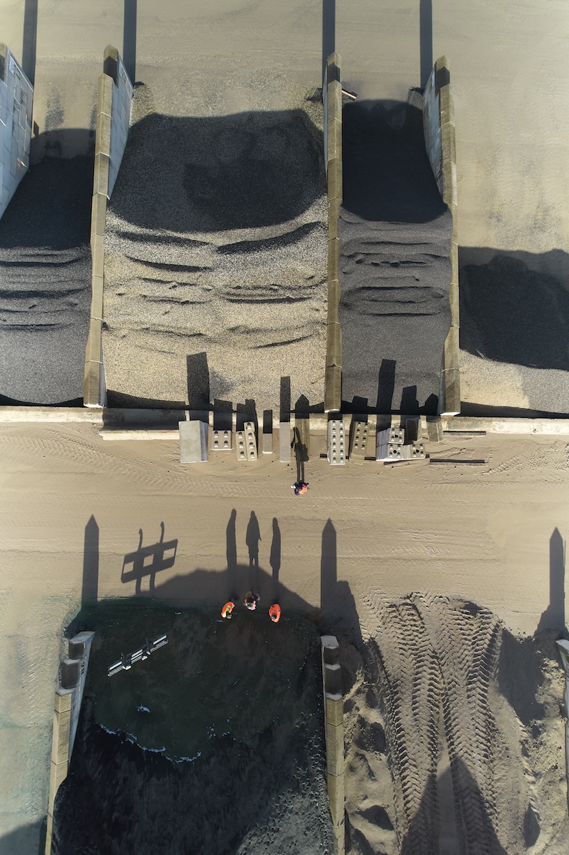 Luftaufnahme einer Baustelle mit großen Silos oder Tanks, an deren Fuß Bauarbeiter und Miniaturmodelle von Hochhäusern zu sehen sind. Im Vordergrund werfen Personen lange Schatten auf den sandigen Boden mit Reifenspuren.