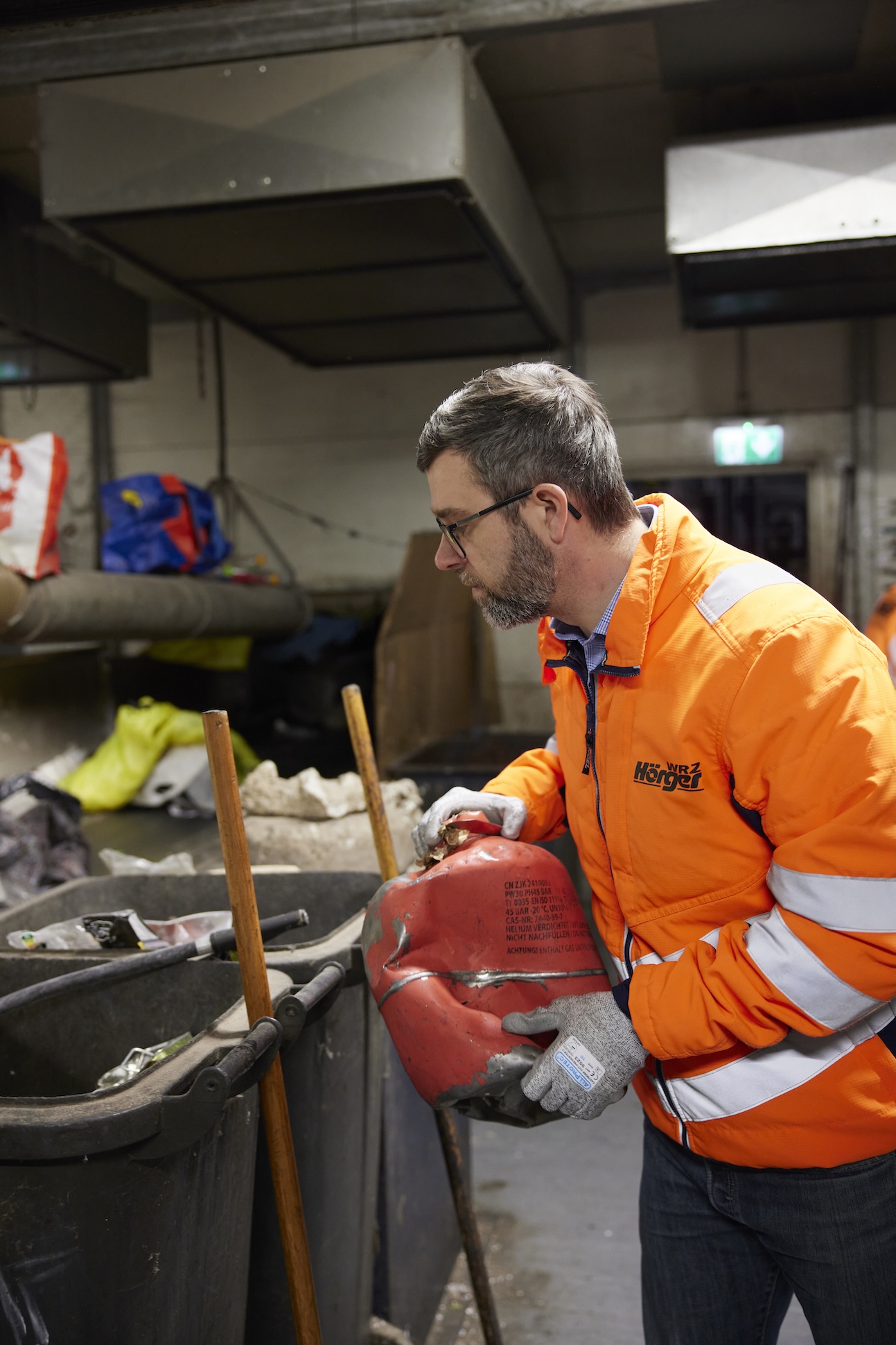 Mann mit Brille in orange-reflektierender Warnschutzjacke und Arbeitshandschuhen hält einen roten Feuerlöscher über einer grauen Abfalltonne in einer Recyclinganlage. Im Hintergrund sind Förderbänder und Sortieranlagen zu sehen.