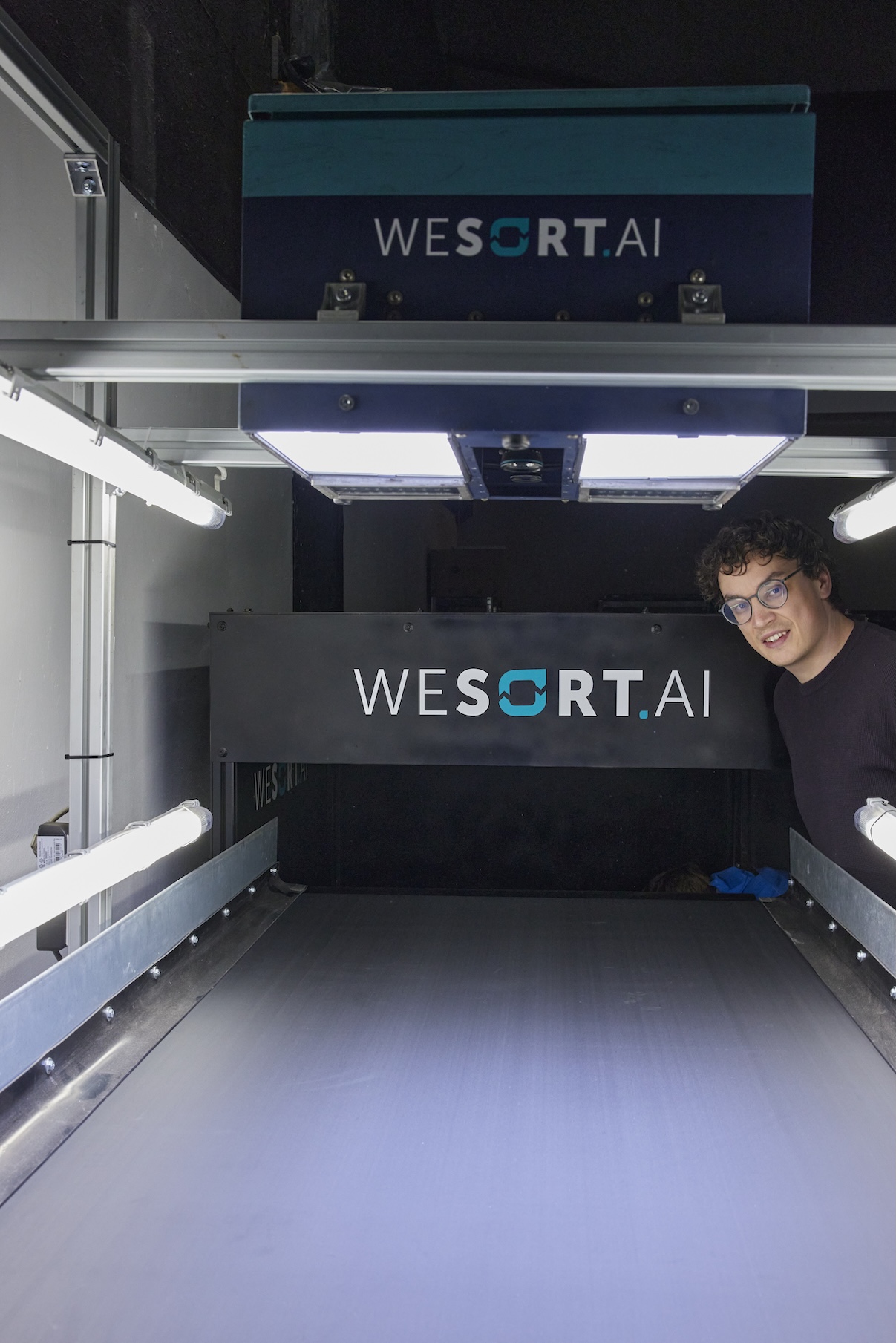 Junger Mann mit Brille und dunklem T-Shirt steht rechts neben einer industriellen Sortiermaschine mit der Aufschrift "WESORT.AI". Die Maschine hat ein Förderband und ist in einem Hallenraum mit Neonbeleuchtung installiert.