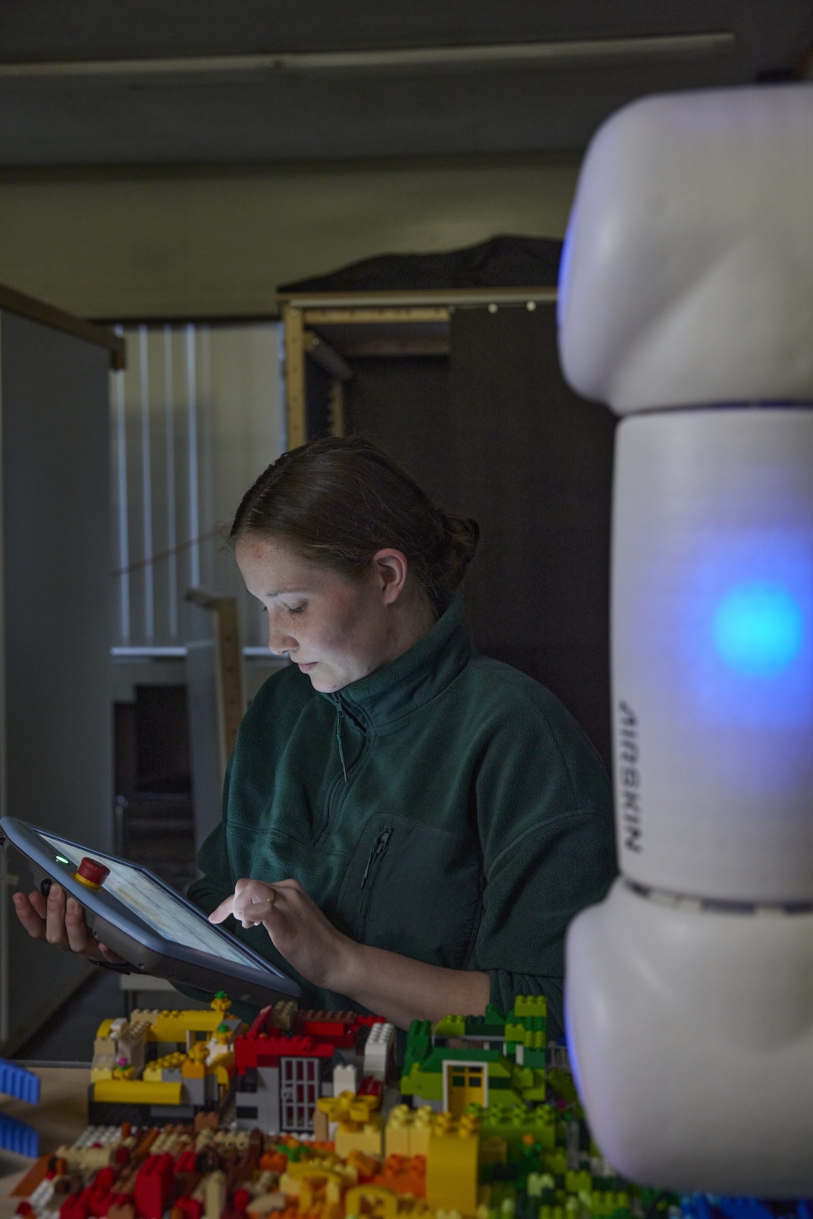 Eine junge Frau in grünem Pullover bedient ein Tablet neben einem weißen Roboter, vor ihr liegt buntes Lego-Spielzeug auf dem Tisch. Die Szene zeigt die Interaktion zwischen Mensch und Maschine in einer Arbeits- oder Forschungsumgebung.