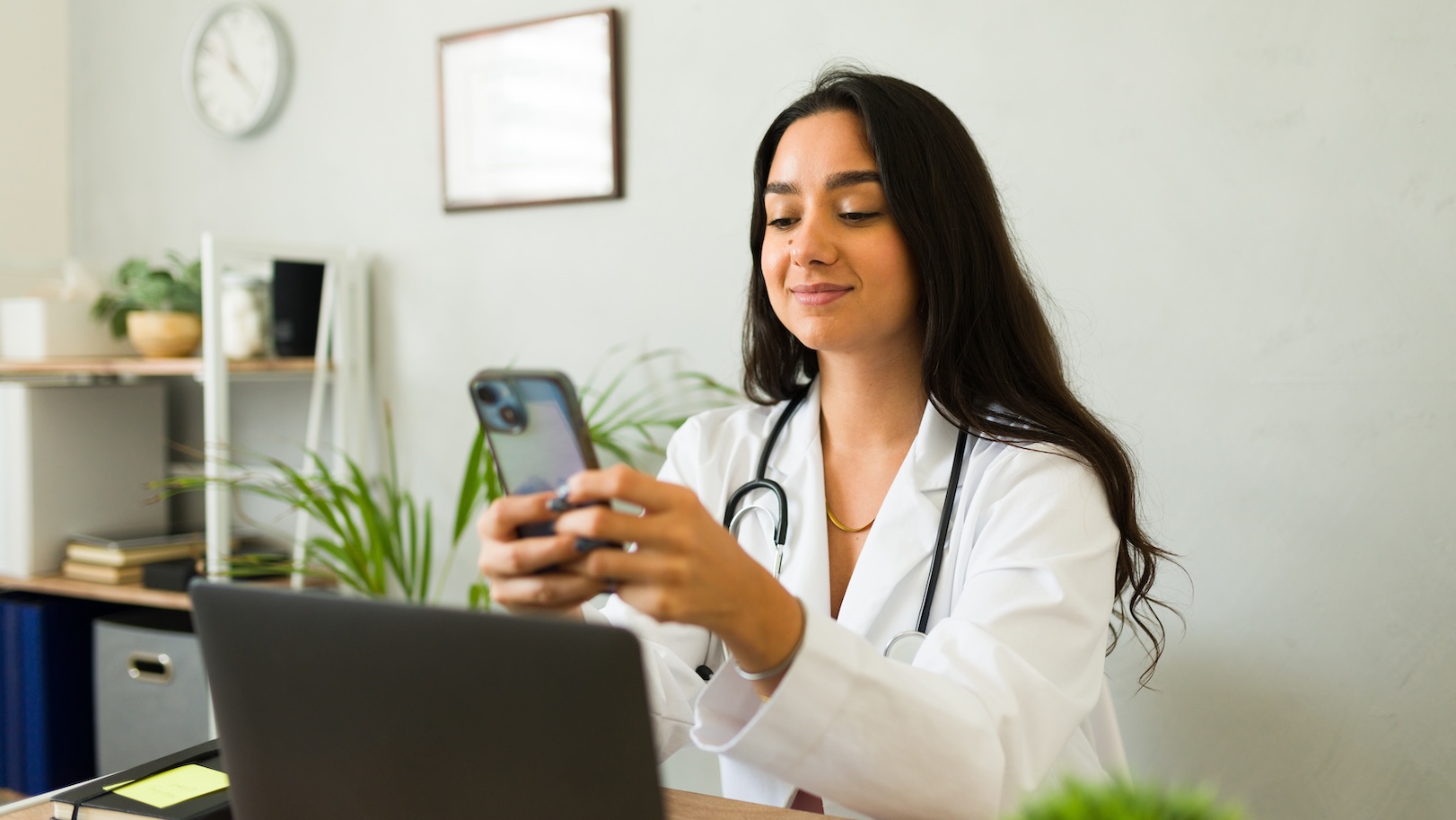 Zu sehen ist eine Ärztin im weißen Kittel mit Stethoskop sitzt an einem Schreibtisch und schaut lächelnd auf ihr Smartphone. Im Hintergrund sind ein Laptop, Regale mit Pflanzen und eine Wanduhr zu sehen.