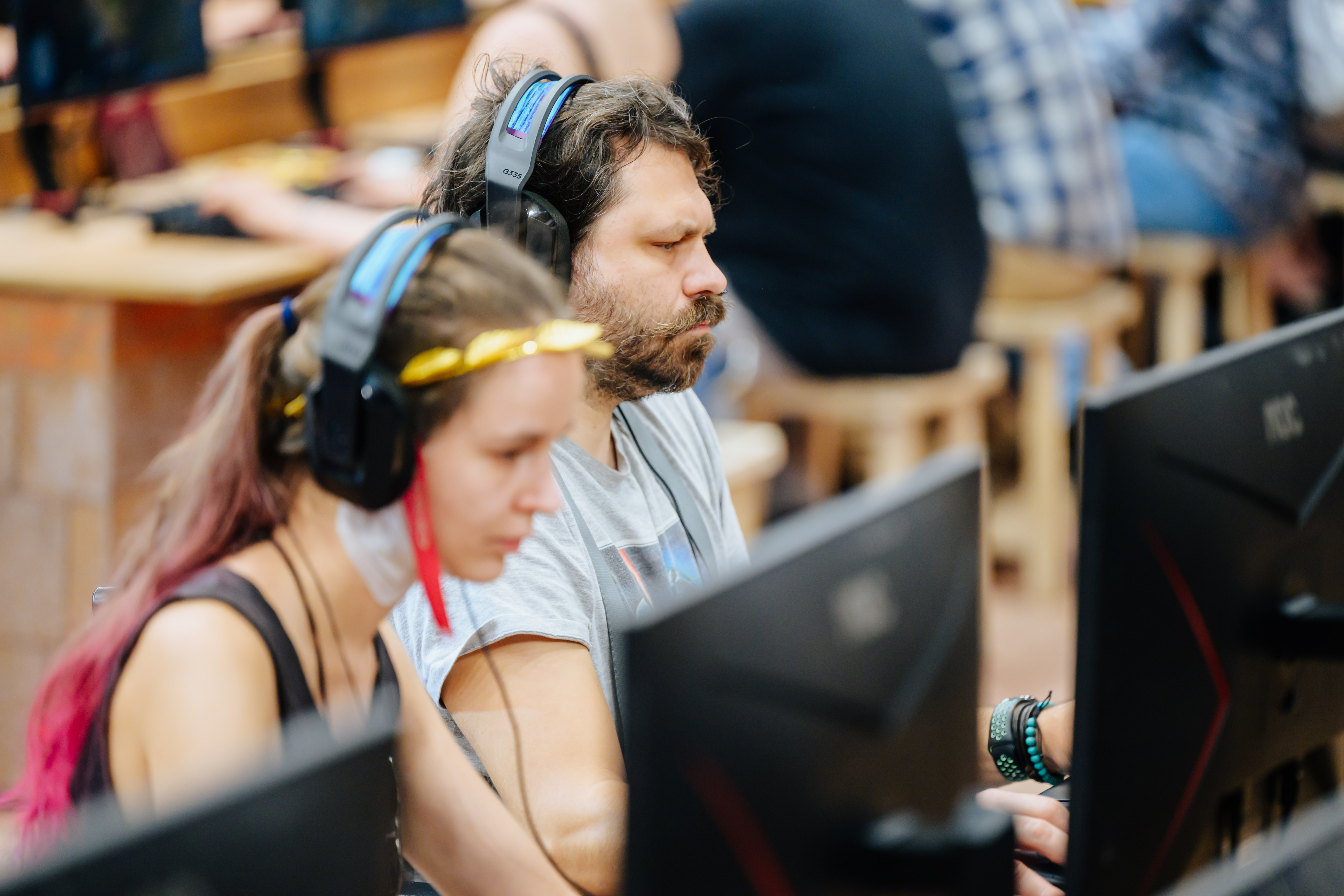 Zwei Gamer mit Headsets sitzen konzentriert vor Computerbildschirmen bei einem E-Sport-Event. Im Vordergrund eine Frau mit pinken Haaren, dahinter ein bärtiger Mann, beide fokussiert auf ihre Spiele.