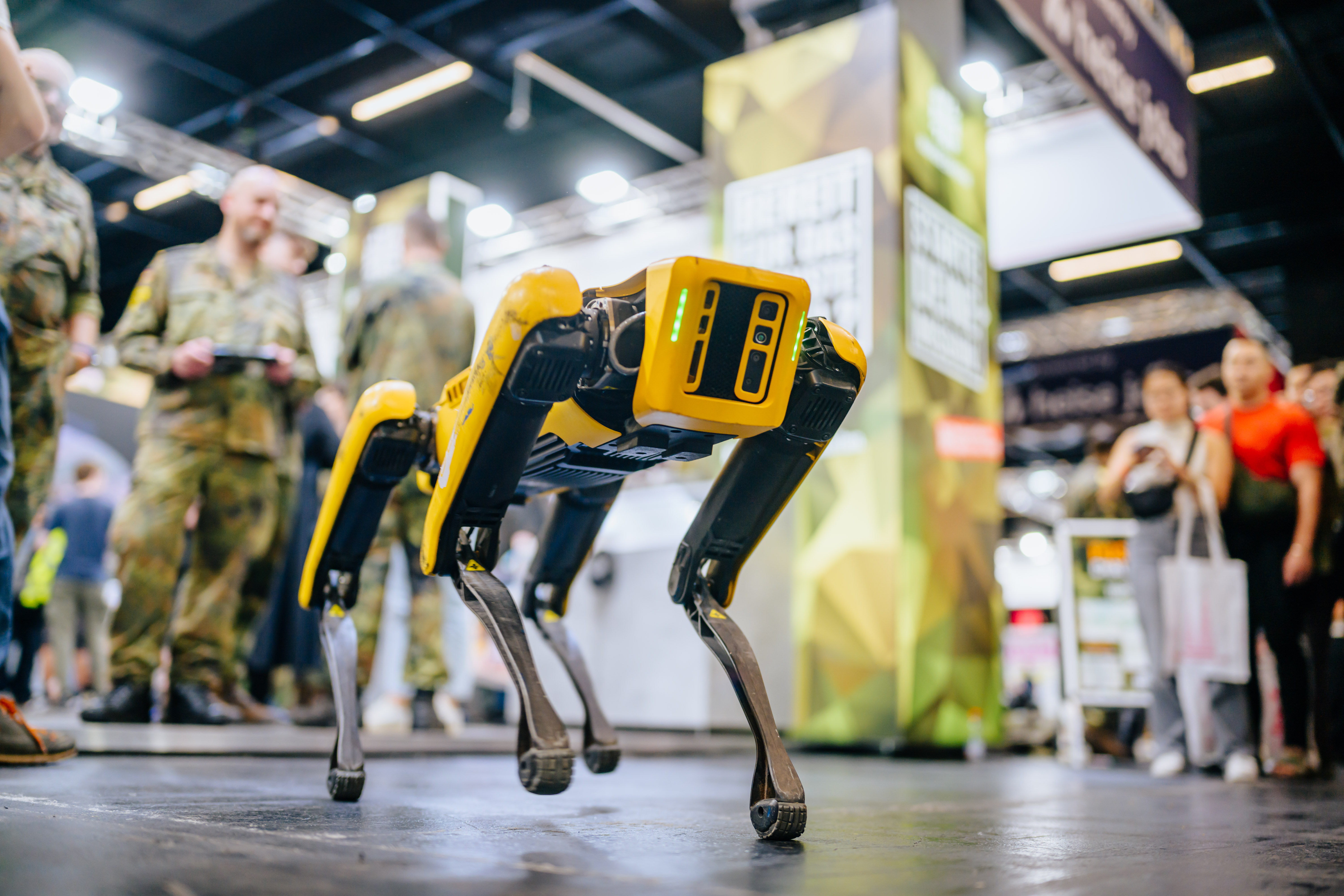 Ein vierbeiniger Roboter in Gelb und Schwarz steht auf einer Messefläche, während Besucher in Militäruniformen und Zivilkleidung die Demonstration beobachten. Der hundeähnliche Roboter präsentiert moderne Robotik-Technologie auf einer Fachmesse.