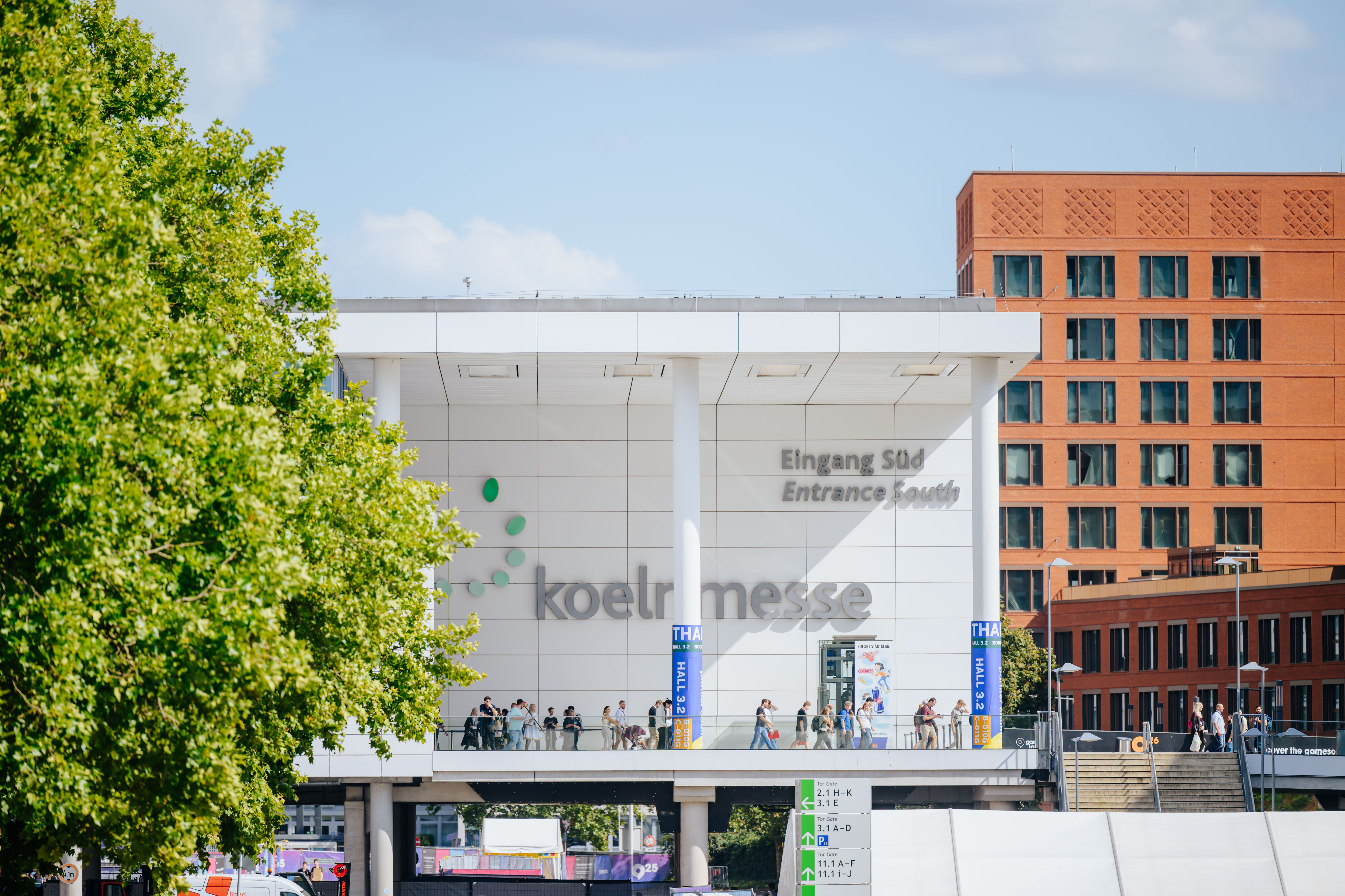 Außenansicht der Koelnmesse mit dem Südeingang, einer überdachten Brücke mit Besuchern und einem modernen orangefarbenen Gebäude im Hintergrund. Im Vordergrund steht ein grüner Baum, Wegweiser zeigen zu verschiedenen Messehallen.