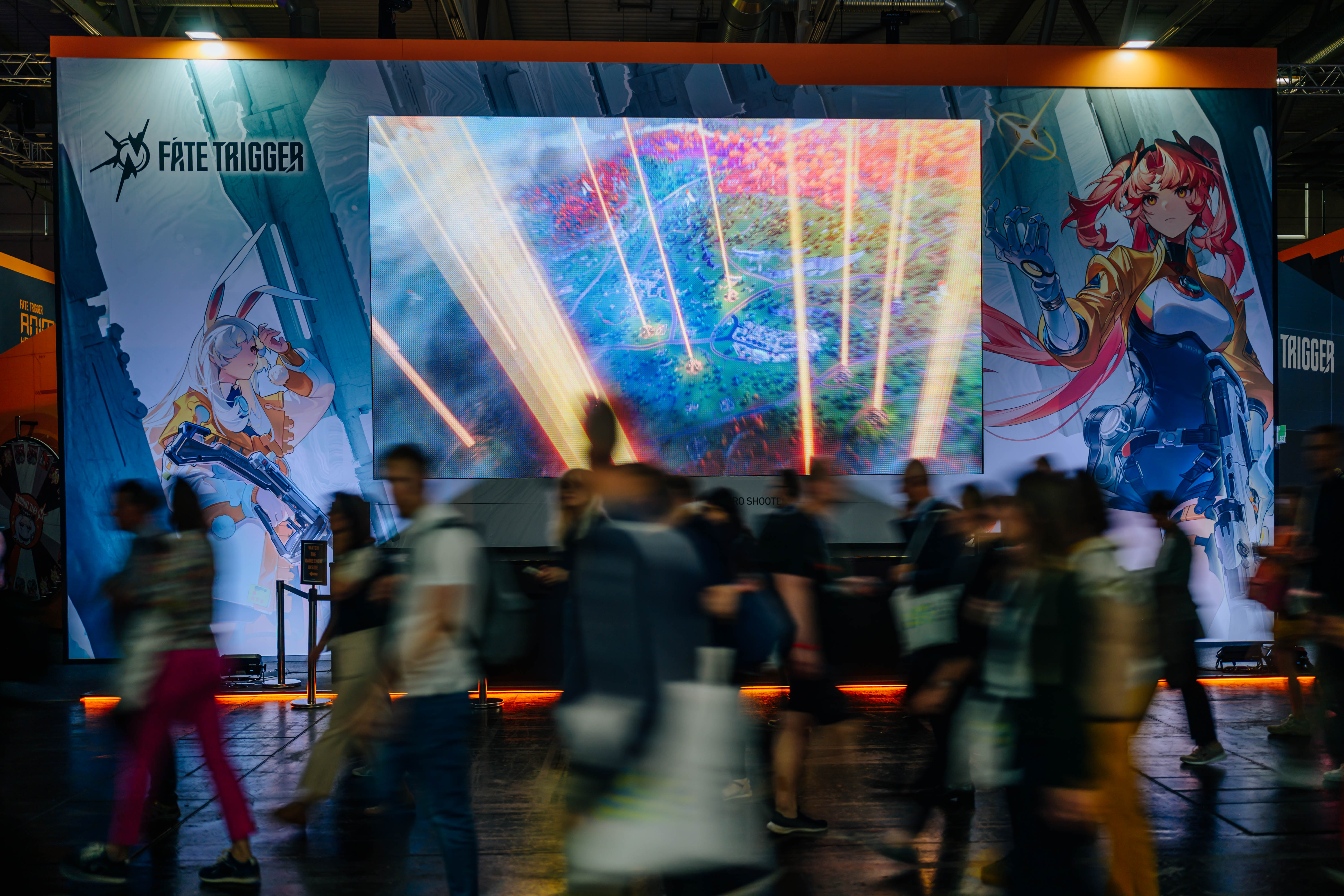 Großer beleuchteter Messestand für das Videospiel "Fate Trigger" mit buntem LED-Display und Anime-Charakteren an den Seiten. Verschwommene Messebesucher bewegen sich im Vordergrund an dem aufwendig gestalteten Gaming-Stand vorbei.