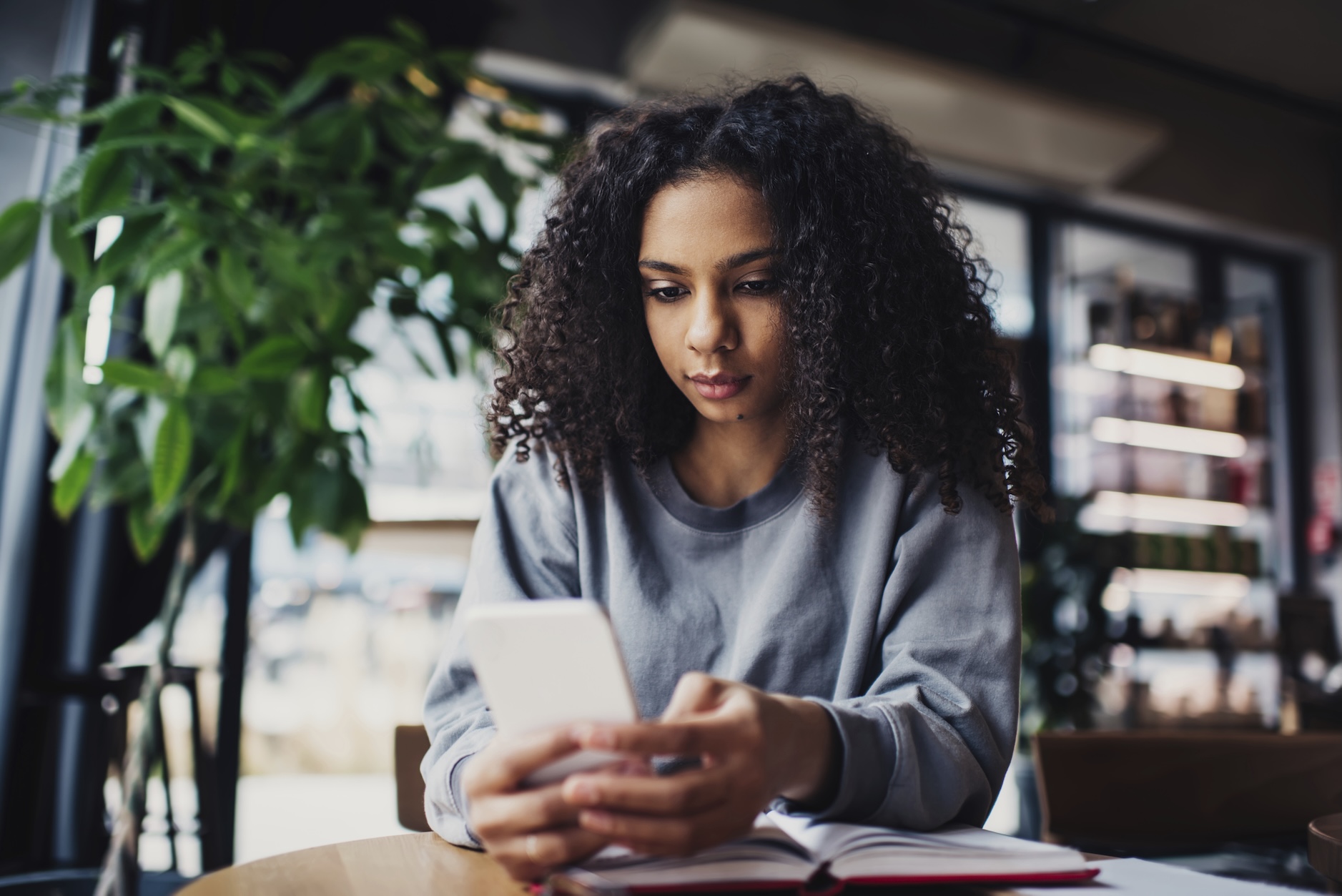 Eine junge Frau mit lockigem Haar sitzt in einem Café und blickt auf ihr Smartphone. Vor ihr liegt ein geöffnetes Notizbuch auf dem Tisch. Im Hintergrund sind Pflanzen und Regale zu sehen.
