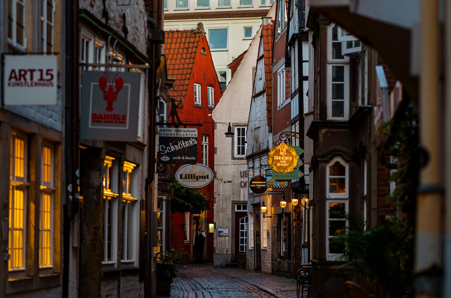 Eine schmale, kopfsteingepflasterte Gasse im Bremer Schnoor-Viertel, gesäumt von historischen Gebäuden mit leuchtenden Restaurant- und Geschäftsschildern bei Dämmerung.