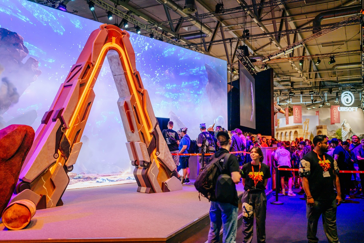 Eine große, orange beleuchtete Spielzeug-Spitzhacke steht als Ausstellungsstück auf einer Gaming-Messe, während Besucher vor einer Videowand mit Fantasy-Landschaft vorbeigehen. Im Hintergrund sind Messestände verschiedener Aussteller wie Ubisoft zu sehen.