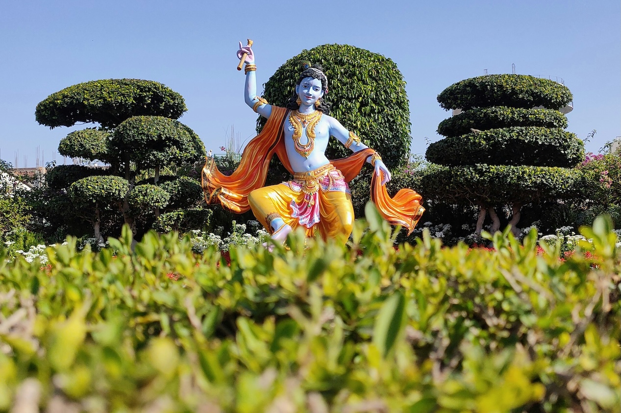 Statue des hinduistischen Gottes Krishna mit blauer Haut, goldgelber Kleidung und orangefarbenem Umhang in einem kunstvoll angelegten Garten mit geschnittenen Bäumen unter blauem Himmel.