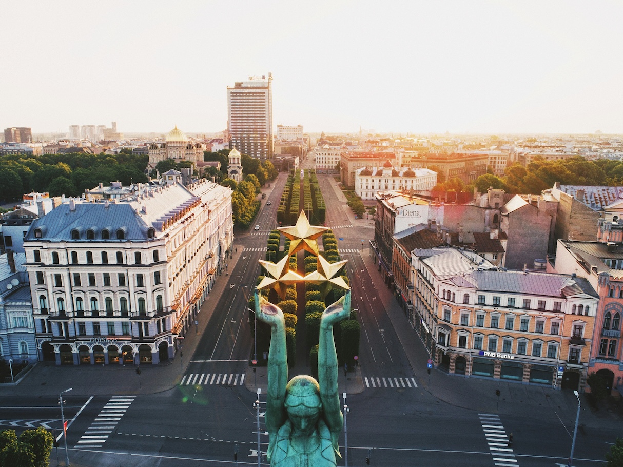 Luftaufnahme des Freiheitsplatzes in Riga mit breiter, baumgesäumter Allee und zentralem Monument mit goldenen Sternen, umgeben von historischen Gebäuden mit klassischer Architektur. Im Hintergrund ragt ein moderner Hochhausturm über die traditionelle Stadtsilhouette hinaus.