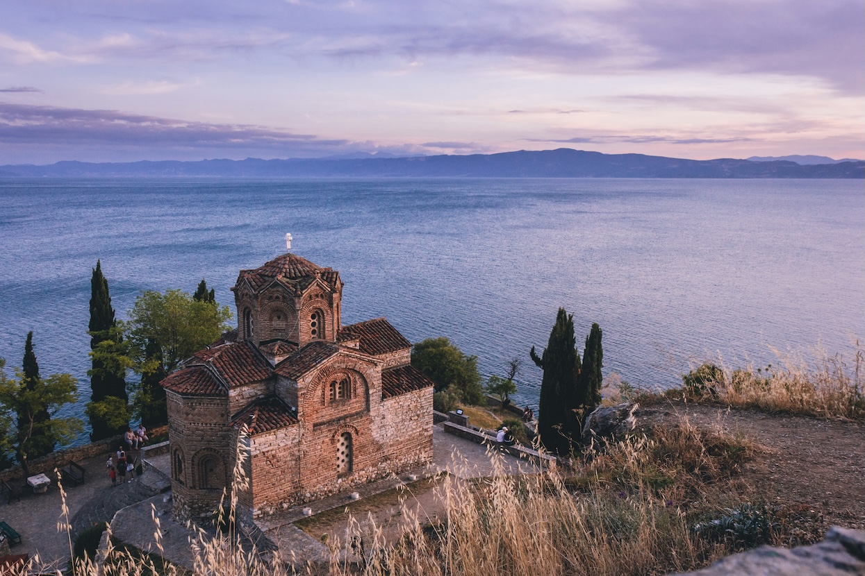 Eine byzantinische Backsteinkirche mit roten Ziegeldächern steht malerisch auf einer Klippe am Ufer eines großen Sees, umgeben von Zypressen im Abendlicht. Im Hintergrund erstreckt sich der See bis zu fernen Bergen unter einem bewölkten Himmel in Rosa- und Blautönen.