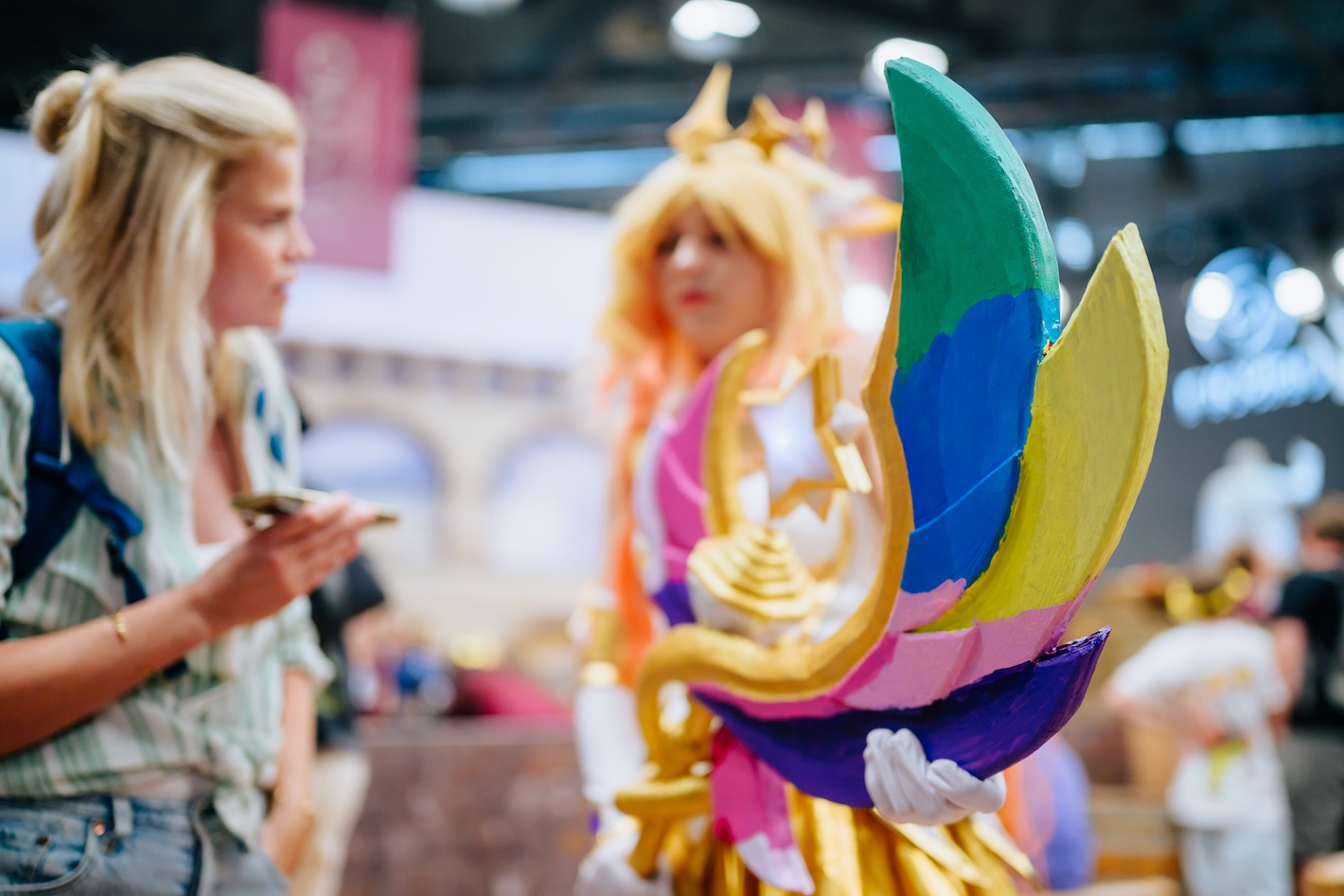 Eine goldene Krone mit bunten Federn in Grün, Blau, Gelb, Rosa und Lila steht im Vordergrund, dahinter sind unscharf Cosplay-Besucherinnen in Kostümen zu sehen. Die Szene wurde auf einer Convention oder Messe aufgenommen.
