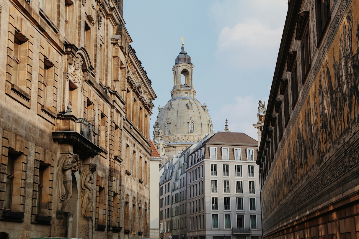 Blick durch eine enge Altstadtgasse in Dresden auf die barocke Kuppel der Frauenkirche, flankiert von historischen Sandsteingebäuden. Rechts ist die Fürstenzugmauer mit ihrem berühmten Porzellanbild-Fries zu sehen.
