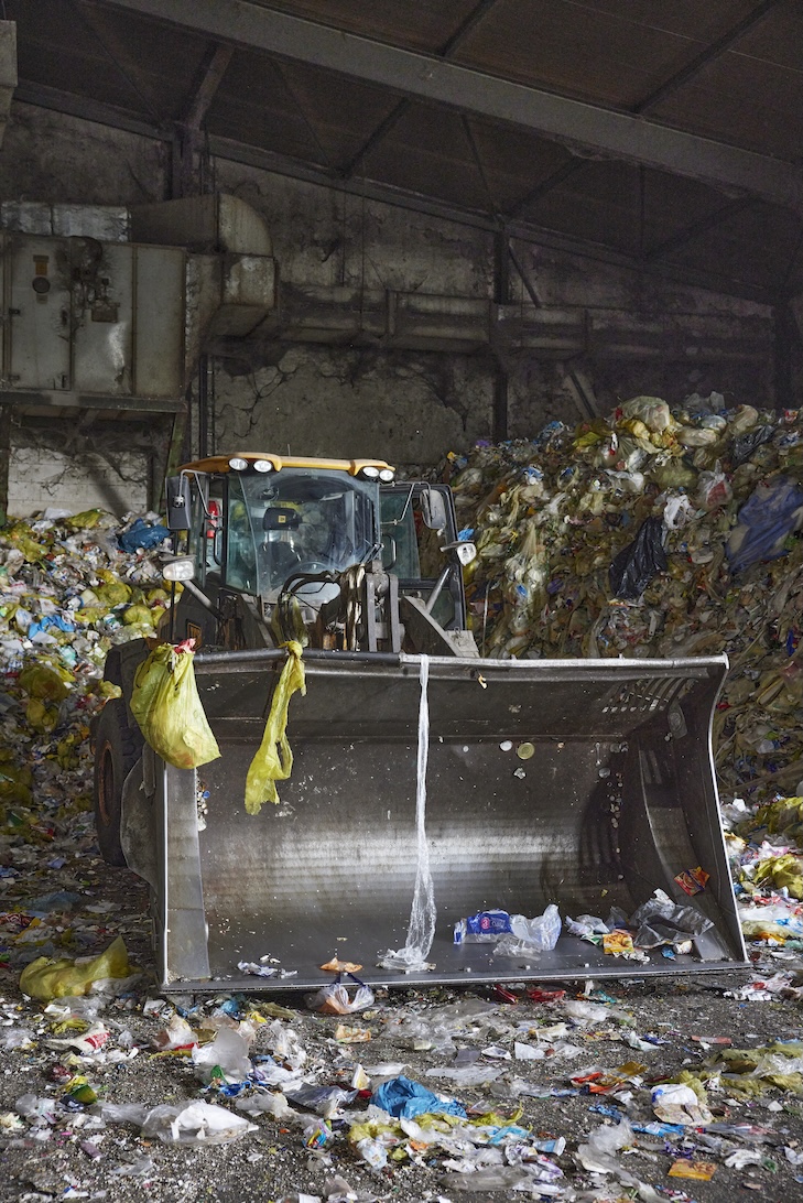 Ein Radlader mit großer Schaufel steht in einer Abfallhalle, umgeben von großen Müllhaufen zu beiden Seiten. An der Schaufel hängen gelbe Müllsäcke, und der Boden ist mit verschiedenen Abfällen bedeckt.Wiederholen