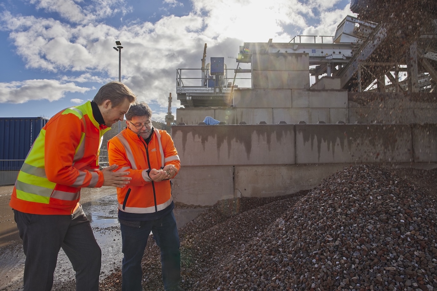 Zwei Personen in orangefarbenen Warnwesten stehen auf einer Baustelle und schauen gemeinsam auf ein Mobilgerät. Im Hintergrund sind Betonwände, Baumaschinen und ein großer Schutthaufen zu sehen.
