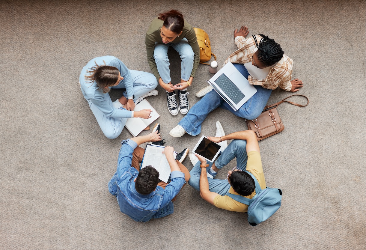 Luftaufnahme von sechs jungen Menschen, die im Kreis auf einem Betonboden sitzen und gemeinsam arbeiten. Sie haben Laptops, Tablets, Notizbücher und Taschen um sich verteilt.