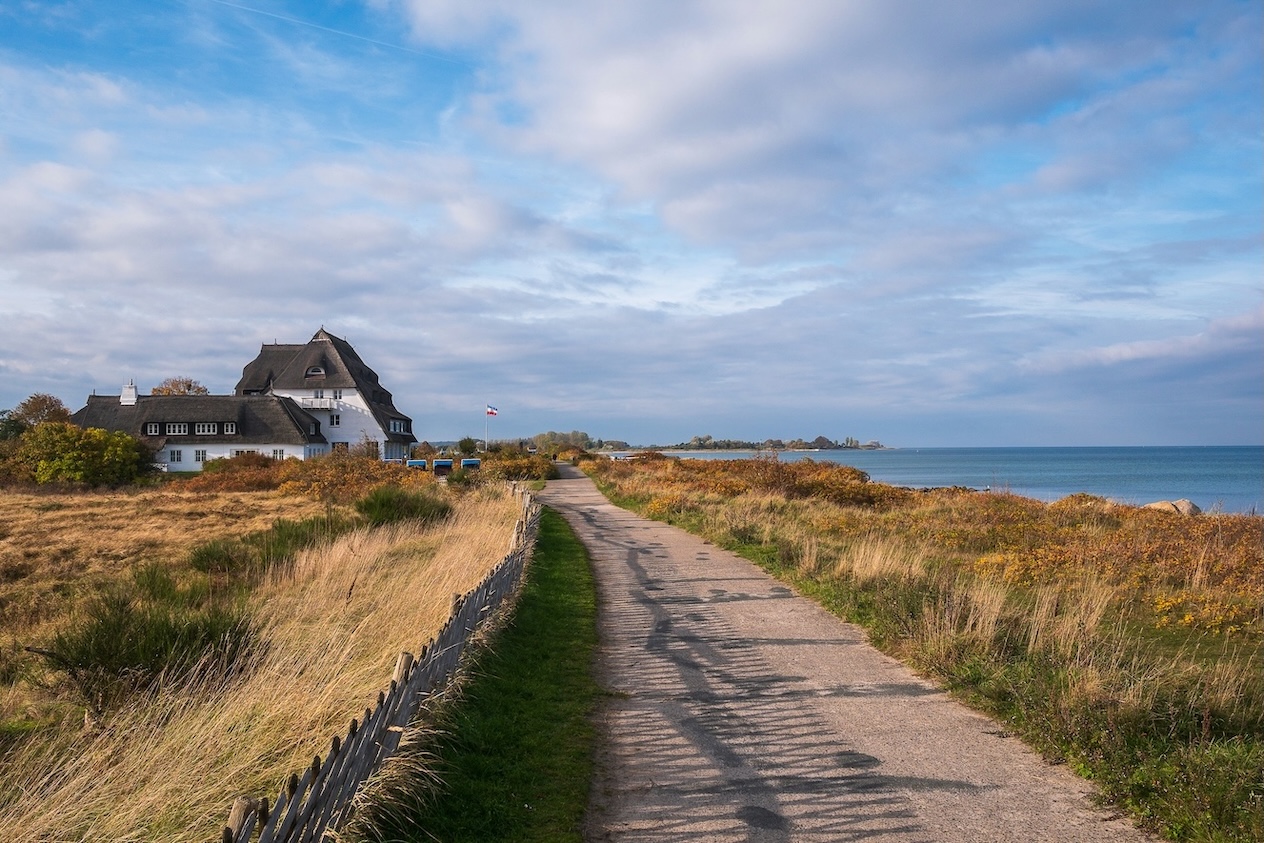 Ein sandiger Weg führt entlang einer Küstenlandschaft mit trockenem Gras und Büschen, rechts das Meer und links ein weißes Haus mit Reetdach unter einem bewölkten Himmel.