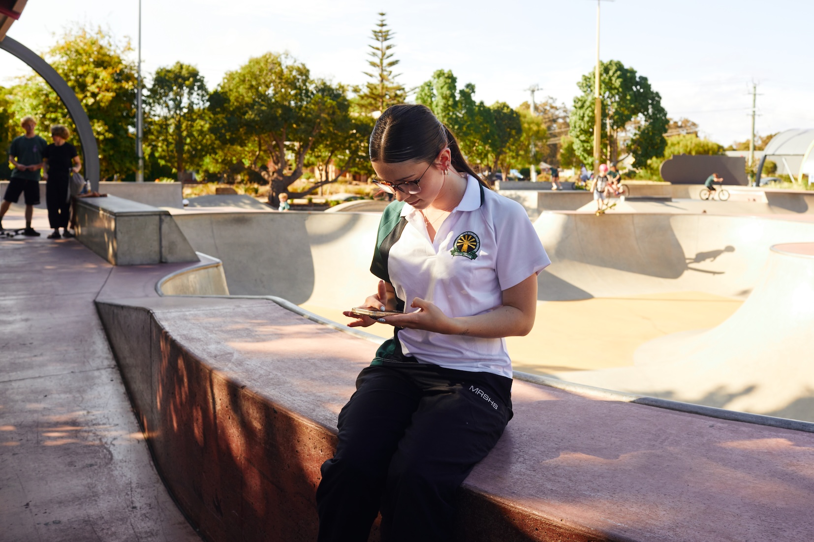 Eine Jugendliche in Schuluniform sitzt an einem Skatepark und schaut auf ihr Smartphone. Im Hintergrund sind weitere Jugendliche zu sehen.