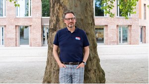 Eckart von Hirschhausen, ein Mann mittleren Alters, steht vor einem großen Baum. Er trägt eine Brille, ein dunkelblaues Polohemd mit einem kleinen Logo auf der linken Brust und eine karierte Hose. Seine Hände sind in den Hosentaschen. Im Hintergrund ist ein modernes Gebäude mit großen Fenstern und einer Fassade aus roten Ziegelsteinen zu sehen. Der Mann wirkt entspannt und freundlich. Die Szene spielt sich wahrscheinlich in einem urbanen Umfeld ab.