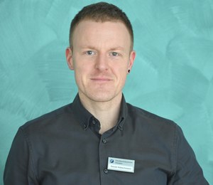 Ein Mann mit kurzem Haar und Ohrring trägt ein dunkles Hemd mit Namensschild und blickt Freundlich in die Kamera. Der Hintergrund ist in einem hellen Türkiston gehalten.