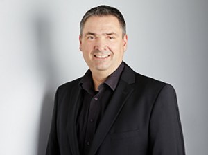 Porträt von Ole Wintermann, in einem schwarzen Anzug mit dunklem Hemd, der vor einem neutralen Hintergrund steht.